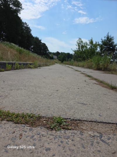 A slightly tilted, upward-angle perspective shot captures a concrete pathway running diagonally into the distance. The pathway is bordered on the left by sparse, dry grass and dirt, with a clear demarcation of the concrete's edge. On the right, the pathway is met by a more substantial strip of green grass and then a dense line of dark green trees. Beyond the trees, a bright blue sky with scattered white clouds dominates the upper portion of the frame. The light suggests it is daytime. A faint, dark line of foliage or a low-lying structure is visible along the edge of the pathway further in the distance, on the right side. The "Galaxy S25 Ultra" watermark is visible in white text in the upper left corner. The scene depicts an outdoor environment, possibly a park or a rural road in Belarus, with no people or animals visible. The focus is on the pathway and the natural surroundings.