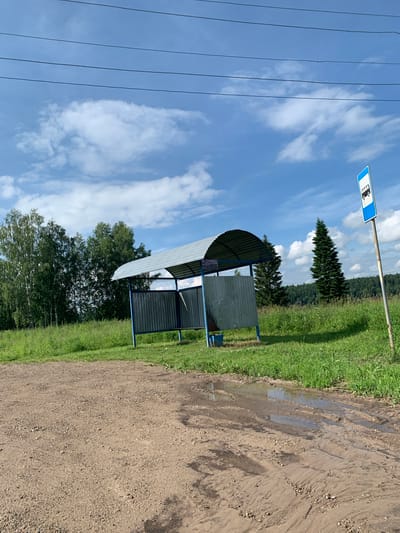 This is a medium-angle, eye-level, outdoor, daytime, photograph taken on a muddy, dirt road in Barsai, Russia. The scene depicts a rural bus stop with a simple, open-sided shelter. The shelter has a corrugated metal roof and green metal walls, supported by blue metal poles. A blue bucket sits on the ground beneath the shelter. To the right of the shelter, a blue and white bus stop sign is mounted on a pole. The background features a grassy field with a line of deciduous trees on the left and coniferous trees on the right. The sky is blue with scattered white clouds, and power lines are visible in the upper portion of the frame. The foreground is a muddy road with tire tracks and puddles of water.
