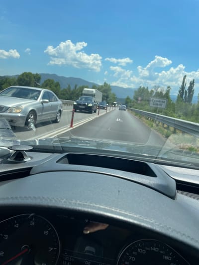 This media file captures a view from inside a car driving on a highway, likely in Bulgaria, given the town name "Simitli" (Симитли) visible on a road sign. The scene is set on a sunny day with a bright blue sky dotted with fluffy white clouds. Lush green trees and mountains form the backdrop.
The primary subjects are the vehicles on the road. The viewpoint is from the driver's perspective, showing the car's dashboard and steering wheel on the right side. A digital display on the dashboard shows the current speed as 57 km/h, along with the outside temperature (+33.5°C) and the time (13:42). Traffic appears to be moving, with several cars and a truck visible ahead. A silver Mercedes-Benz is prominently seen in the foreground from the driver's side.
Notable details include the road sign indicating "Simitli," suggesting the car is either approaching or leaving the town. The weather is clearly warm and sunny, indicated by the high temperature and the clear blue sky. The overall mood appears to be one of travel on a pleasant day. The dashboard instrumentation, including a speedometer that goes up to 200, is also visible. The quality of the image suggests it was taken with a smartphone.