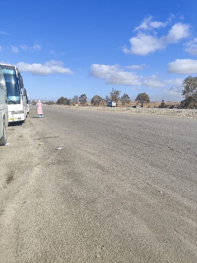 A long stretch of paved road fills the foreground, leading towards a distant horizon under a bright blue sky with scattered white clouds. To the left, several large white buses are parked. A lone figure in a pink robe walks alongside the buses towards the background. Across the road, sparse trees and vegetation line the horizon. Further back, several vehicles are parked.