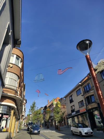 The image captures a street scene in Lessines, Belgium, with buildings lining both sides of the road under a clear blue sky. Suspended from wires across the street are several decorative elements: a large blue top hat, a red pipe, and a pink umbrella, with a smaller red oval shape visible in the distance. A tall lamppost with a reflective surface stands on the right side of the frame, partially obscuring the view of the sky.

On the left, a curved building with brick and white facade details features several windows and a balcony. A storefront window on the ground floor displays clothing with a "SOLDES" (Sales) sign.

The street itself is paved, with tram tracks visible running down the center. Two cars are present: a dark blue car in the middle of the street and a small white car parked on the right. Several trees with green foliage are planted along the sidewalk, interspersed with other buildings of varying architectural styles and colors. Some of these buildings have storefronts, with one signage reading "GAKO". The overall impression is of a moderately busy street in a European town during daylight hours.