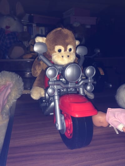 A stuffed monkey is perched on a toy motorcycle. The motorcycle is red and silver with a black tire. The monkey has brown fur and black eyes. The background is dark and blurry, with shelves of toys visible. The scene appears to be indoors, possibly in a child's room. The location of Tutaev, Russia is not depicted in the image.