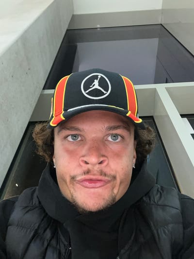 A man wearing a black hooded jacket and a black baseball cap with a white "Jumpman" logo and orange and yellow trim is facing the camera. He has light skin, short brown hair, a goatee, and gray eyes. The background consists of a white wall, a gray ledge, and a dark, reflective surface that may be a window. The location may be Ivry-sur-Seine, France.