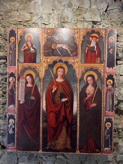 This is a detailed altarpiece depicting several religious figures, painted in a Gothic style with gold leaf accents. The altarpiece is divided into multiple panels.
In the top row, from left to right, are: a standing monk with a halo and staff; Saint John the Baptist holding a lamb and a book; a depiction of the Lamentation of Christ, with Christ's body being held by figures in dark robes; and a man in red robes with a red hat, holding an open book, with a lion at his feet. To the right of the man in red is another standing monk in dark robes.
The middle and bottom rows feature several standing female figures, all with halos. The central figure is a woman in a red robe, holding a book and a palm frond. To her left is a woman in a dark robe, holding a palm frond and a tower. To her right is another woman in a dark robe, holding a palm frond and with exposed breasts, suggesting she is Saint Agatha. Flanking these central figures are smaller panels on the sides. On the left side, a figure in episcopal vestments holds a crozier, and below him, a nun holds a white flower. On the right side, a monk holds a book, and below him, another nun holds a white flower.
The altarpiece is hung on a stone wall, suggesting it is displayed in a historical building or museum. The style of the painting, particularly the gold halos and the depiction of the figures, is characteristic of medieval religious art.
This altarpiece is located in the Church of Santa Croce in Dolceacqua, Italy.