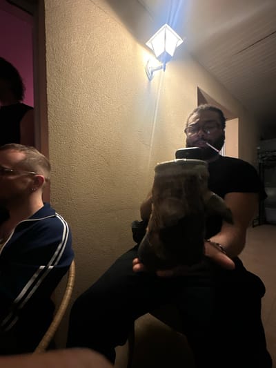 The image shows an indoor scene with two people and what appears to be a small animal. The man in the foreground has blond hair and wears a blue shirt with white stripes. The other man in the background wears glasses, a black shirt, and dark pants. He is holding a furry animal. The image is taken from a low angle, looking up. On the wall behind them is a light fixture. The background suggests an indoor or enclosed outdoor space with a textured wall. The location is Montreuil, France.
