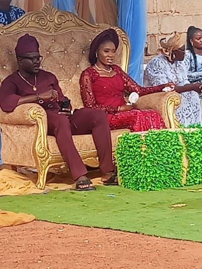 This image captures an outdoor event in Afaka, Nigeria, featuring two individuals prominently seated on ornate golden, tufted chairs. The male on the left, with dark skin, wears a maroon traditional ensemble comprising a hat, long-sleeved top, and trousers, complemented by dark-framed glasses, a gold chain with a circular pendant, and a bracelet on his left wrist. He holds a dark-colored smartphone in his left hand and wears black slippers. The female on the right, also with dark skin, wears a long-sleeved red dress with sequined embellishments, a matching headwrap, a white pearl necklace, and earrings. She holds a small, white, circular object in her right hand and wears black footwear. Her gaze is directed towards her left (viewer's right).

The chairs rest on a green artificial turf, which meets a reddish-brown dirt ground in the immediate foreground. Yellow fabric is draped beneath the chairs. In front of the chairs, two square blocks covered in artificial green foliage are present. The background features blurred figures, including an individual with a patterned light-colored headwrap and another in a light blue top. A light-colored stone or brick wall structure is visible, along with blue fabric drapery further left. The scene is illuminated by bright natural daylight.