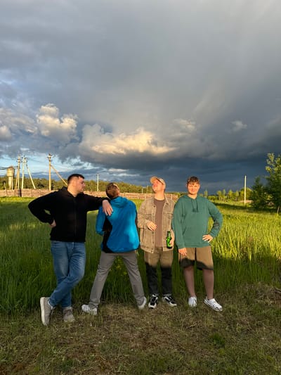 The image depicts four men lying in a grassy field in what appears to be a rural area. In the background, there are utility poles and a line of trees. The sky is dramatic, with a mix of dark, stormy clouds and patches of lighter, sunlit clouds.
From the foreground to the background:
- The first man is wearing a black long-sleeved shirt, blue jeans, and grey sneakers. He is positioned with his arm raised towards the sky and another person behind him.
- The second man is wearing a blue jacket and grey pants, lying on his back.
- The third man is wearing a brown jacket over a dark shirt and dark pants, lying on his back and appears to be holding a green bottle.
- The fourth man is wearing a green hoodie and brown shorts with white clogs.
The grassy field appears to be unkempt, with varying lengths of grass. The overall impression is one of a casual outdoor setting. The location could be Vysokinichi, Russia, based on the request.