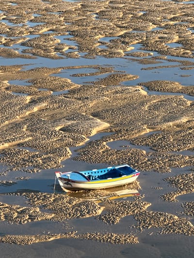 A boat is stranded in the sand in Vila Real de Santo António, Portugal. The sand is rippled and wet, reflecting the sky. The boat is blue and white with yellow trim, resting on two parallel rails.