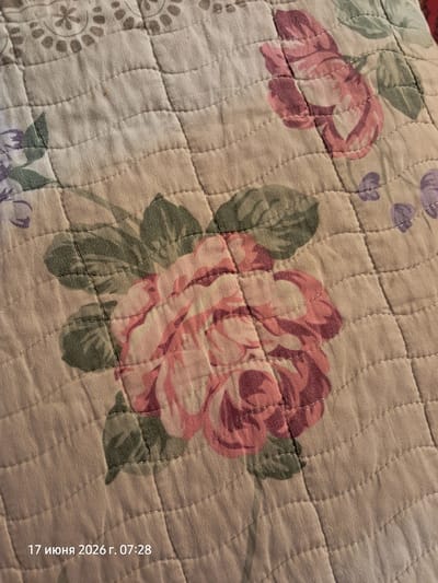 The image shows a close-up view of a quilted fabric with a floral pattern. The pattern features large pink and red roses with green leaves. The stitching of the quilt creates a grid-like texture. In the bottom left corner, text reads "17 июня 2026 г. 07:28" which translates to "June 17, 2026 at 07:28". There are no people or recognizable landmarks visible in the image. The location in Gumbaz, Uzbekistan is not discernible from the image itself.