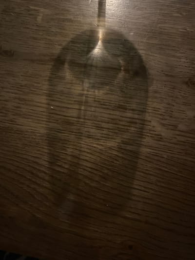 The image depicts a wine glass lying horizontally on a wooden surface. The wood has a prominent grain pattern. The glass is mostly transparent and creates a darker shadow around its outline, suggesting an empty glass or one containing a clear liquid. The glass's stem is visible to the left. There is no explicit information connecting this image to the city of Rehovot, Israel. No people, actions, or interactions are discernible. The scene is indoors with an unknown light source casting the shadow.