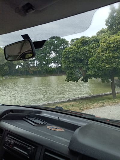 This media file provides a view from inside a vehicle, looking out through the windshield. The scene depicts a calm body of water, likely a pond or small lake, bordered by a stone embankment and lush green trees. In the foreground, the dashboard of the car is visible, with a mobile phone and a small, circular metal object placed on it. A car's rearview mirror is also in view, reflecting the sky and trees.
The weather appears to be overcast, suggesting a cloudy day. The time of day is not explicitly clear but seems to be daytime. The location is identified as Svémyslice, Czechia.
There is no discernible activity or event taking place; the scene is static and peaceful. Notable details include the texture of the water, the dense foliage of the trees, and the arrangement of objects on the dashboard. The overall impression is one of a quiet moment observed from the comfort of a vehicle, possibly during a scenic drive or a stop to enjoy the view. No visible text is present in the image.