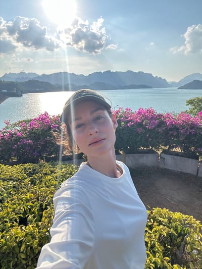 The image shows a young woman taking a selfie in Ban Chiao Lan, Thailand. She is wearing a white shirt and a cap. The background includes a body of water, likely the Cheow Lan Lake, with mountains in the distance and flowering plants (possibly bougainvillea) and other greenery in the foreground. The sky is sunny with some clouds. The photo is taken during the daytime, with the sun shining brightly.