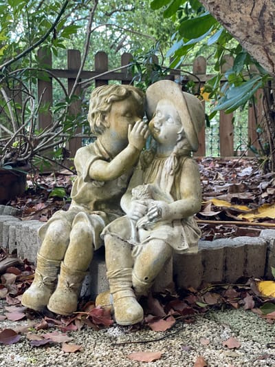 The image shows a garden sculpture of two children. One child wears a hat and appears to be lying on top of the other. The sculpture is located on a low brick structure, possibly steps, surrounded by leaves and greenery. A wooden fence is visible in the background. There is no information available in the image to confirm the city of Clearwater, United States of America.
