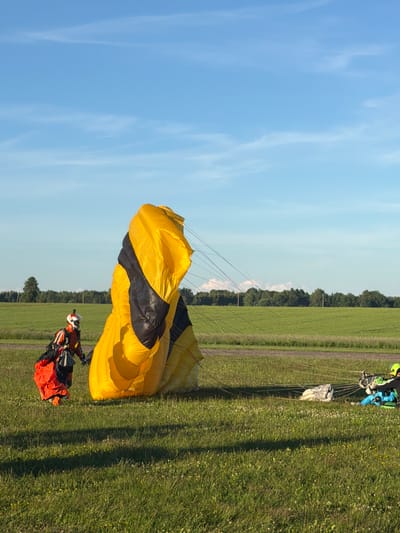 The image captures a bright, sunny day with a clear blue sky and scattered light clouds, set in a grassy field that appears to be an airfield or a large open park, possibly located in or near Limbaži, Latvia. In the foreground, a person dressed in a bright orange and black skydiving suit with a white helmet is seen on the left side of the frame, partially interacting with a large, yellow and black parachute that is partially deployed and resting on the ground. The parachute is the dominant object in the mid-ground. To the right, another person, wearing a blue and green suit and a green helmet, is seated on the ground, seemingly involved with equipment. The ground is covered in green grass with some small white and yellow wildflowers. In the background, a treeline with lush green foliage runs across the horizon, separating the field from the sky. The overall scene suggests an activity related to parachuting or skydiving, possibly post-landing or preparation for a jump.