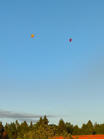 Two hot air balloons, one yellow and one red, are visible in the clear blue sky above a treeline. The yellow balloon is to the left and slightly higher than the red balloon on the right. Below the sky, a dense line of green trees forms a silhouette against the sky. The lower part of the image shows a section of an orange-tiled roof. The scene appears to be in a rural or suburban area, likely during the daytime due to the bright sky and illumination of the trees. The location is identified as Novonikolskoye, Russia.
