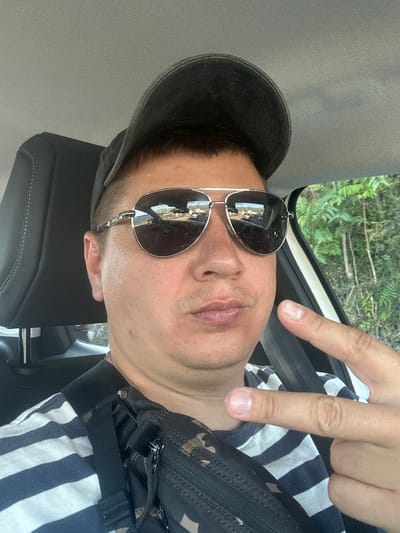 A man is taking a selfie in a car. He is wearing a dark baseball cap, aviator sunglasses, and a striped t-shirt. He has a camouflage fanny pack on. He is making a peace sign with his right hand. In the background, through the car window, there are green trees. The image does not contain any visual information that would suggest a specific location in the city of Budva, Montenegro.