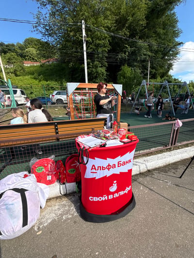 In Prohress, Russia, an outdoor event is taking place near a playground. A red and white branded table displays various items, including pamphlets and promotional materials. Several red backpacks with "Alpha Bank" branding are placed on the ground near the table. A woman with red hair, wearing sunglasses and a black t-shirt, stands behind the table, holding a camera or phone. Children are visible in the background, some playing on the swings and playground equipment. There are cars parked in the background and trees and a grassy hillside on the left side of the image. The ground is paved with asphalt and concrete.