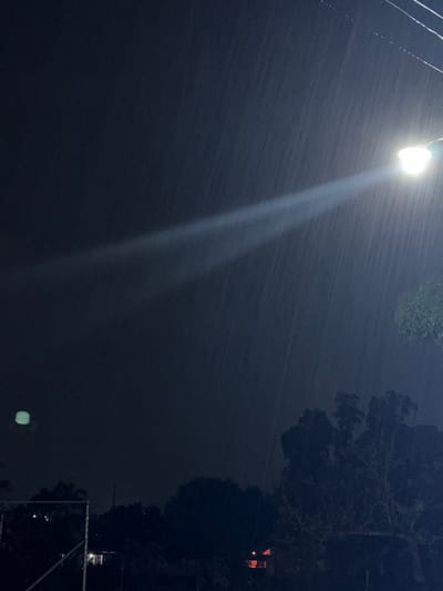 The image depicts a night scene, likely in El Bajío, Venezuela, characterized by heavy rain illuminated by a street lamp. The lamp is positioned at the upper left, casting a bright, white light downwards through the falling rain. To the right, dark foliage suggests trees, with a building visible in the distance exhibiting a lighted window. In the lower right corner, part of a chain-link fence is visible. The overall impression is one of a dark and wet environment, typical of a rainy night. No people are visible.