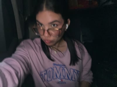 A young female with dark hair and glasses is captured in a close-up shot, wearing a light purple sweatshirt with the "Tommy Jeans" logo. She has on white earbuds. The background is dark and out of focus, offering no discernible details about her surroundings or the specific location within Conuco Viejo, Venezuela.