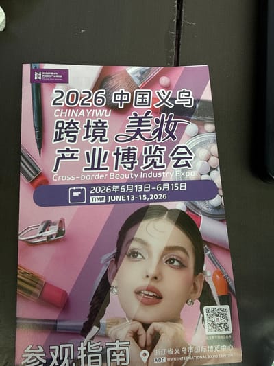 This is a photograph of a promotional flyer for the "2026 China Yiwu Cross-border Beauty Industry Expo." The flyer features a close-up of a young woman with dramatic eye makeup and braided hair, surrounded by various cosmetic products like lipstick, mascara, eyelash curlers, and makeup brushes. The text on the flyer is a mix of Chinese and English, with the event title prominently displayed. Key information includes the event dates: June 13-15, 2026. At the bottom of the flyer, it indicates the location as "ADD YIWU INTERNATIONAL EXPO CENTER," which is in Yiwu City, Jin Hua Shi, China. A QR code is also visible, likely for more information or registration. The flyer is placed on a dark, textured surface, possibly a table.