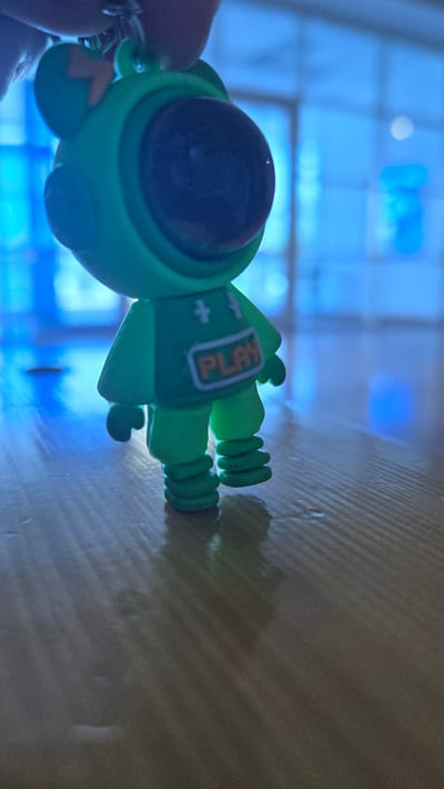 The image displays a close-up, low-angle shot of a green, cartoonish figurine, possibly a keychain charm, standing on a wooden surface. The figurine resembles a frog or a similar amphibian creature, with large, dark eyes and a vest-like garment displaying the word "PLAY" in orange letters. The background is blurred and predominantly blue, suggesting an indoor setting with bright, artificial lighting, possibly through windows. The wooden surface exhibits a visible grain pattern and reflections, indicating it may be slightly damp or glossy. No specific city in Kaka, Turkmenistan is identifiable from the visual information.