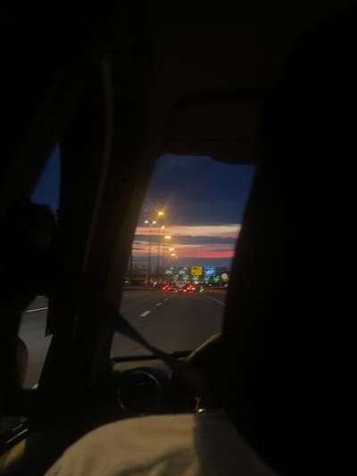The image is a dark, blurry shot taken from inside a car at dusk. Through the car window, a highway is visible with lanes marked by white lines and illuminated by streetlights. Several cars with illuminated taillights are driving on the highway. In the distance, there are traffic signs and overpasses. The sky shows a mix of dark blue and colorful hues of orange and pink, suggesting a sunset or sunrise. The foreground is dominated by the dark interior of the car, including a seatbelt and part of a headrest. The image does not provide any specific details to confirm the location in Pesochny, Russia.