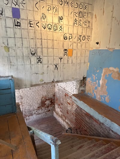 The image depicts a weathered interior space with a prominent wall covered in a grid of squares, some containing black symbols and others left blank. There are streaks of black paint running down the wall, suggesting artistic execution or deterioration. Two small squares, one purple and one orange, are affixed to the grid. To the right, a section of blue-painted wall is visible, with peeling paint revealing lighter material underneath. Handwritten text in black marker reads "Eve Horr" on this blue section.
In the foreground, wooden stairs descend into a lower level. The stairs and surrounding platforms are made of worn, unfinished wood, showing signs of age and use. A light blue wooden door is visible on the left side of the stairs. The lower level walls are constructed of exposed brick, with some sections whitewashed and others showing the raw brick. The overall impression is of an abandoned or neglected space, possibly an art installation or an old industrial building.
The location is identified as the former administrative building of the Yelets Confectionery Factory in Yelets, Russia. This building is known for its artistic transformations, including the wall art visible in the image, created by artist Valery Gryaznov.
