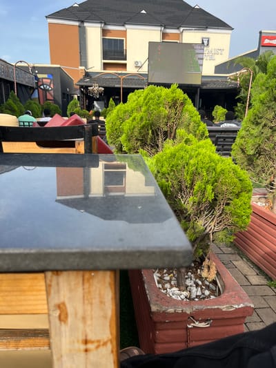 The image depicts an outdoor scene, possibly in the city of Offatedo, Nigeria, given the visible "Skymist Hotel" sign in the background. The foreground is dominated by a black marble tabletop supported by a wooden frame. The reflection on the tabletop reveals a building with a brown and beige facade, likely the Skymist Hotel.
Several green, trimmed bushes are placed in planters and line the area. One planter, made of weathered red concrete and filled with small white stones, sits close to the tabletop. A pair of white electrical wires is visible attached to this planter.
Further into the background, figures of people are barely visible. A large video display screen is placed outside the hotel. The sky is overcast.