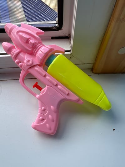 A pink plastic toy water gun is positioned on a white surface. The water gun has a rocket-shaped body and a yellow reservoir. The image is cropped to show the water gun and a hint of a window with a screen in the background. There is no indication of the city of Мариинский, Russia, or any specific urban environment.