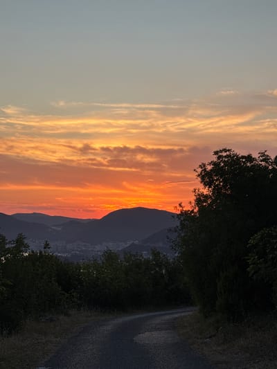The image captures a vibrant sunset over the mountains near Čelobrdo, Montenegro. The sky is ablaze with hues of orange, red, and yellow, with streaks of pink and purple visible in the clouds. Silhouetted against the colorful sky are rolling mountains, with a town nestled in the valley below, faintly illuminated by the fading light. In the foreground, a gravel road winds its way through dense, dark green foliage, suggesting a natural, slightly rugged landscape. The overall scene evokes a sense of tranquility and natural beauty as day transitions into night.