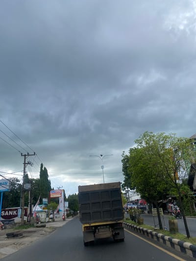 The image depicts a street scene in Sungai Paring, Indonesia, under a cloudy, overcast sky. A yellow dump truck with a black bed is the primary subject, driving away from the camera on the right side of a paved road. The truck has dual exhaust pipes. The road has a distinct black and white striped curb. To the left of the road, a sidewalk area is visible, with stacked tires and various structures including a utility pole with wires. Signs are present, including one for "Alfamart" and another with the text "SANKI". There are trees along the roadside, with one prominent green tree on the right side of the image. A few motorcycles and a three-wheeled vehicle are also visible, suggesting a moderately busy urban or suburban environment. The overall atmosphere is muted due to the cloudy weather.