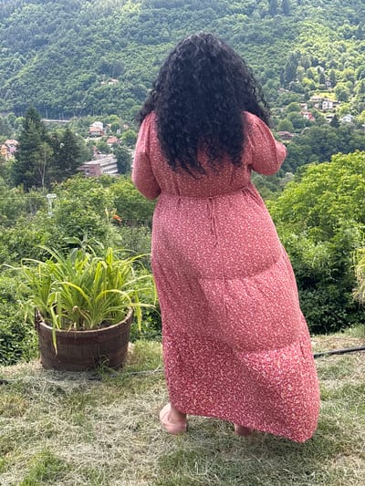 A person is standing in a garden, viewed from behind. They are wearing a long, red, floral-print dress. The person has long, dark, curly hair. They are standing on a grassy area, with a potted plant to their right. In the background, there is a lush green hillside covered with trees and houses, suggesting a rural or suburban location. The lighting suggests it is daytime, and the greenery indicates pleasant weather. The overall scene evokes a feeling of peacefulness and connection with nature.