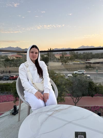 A female individual, appearing in her 20s or 30s, is seated in a light-colored, possibly woven, chair at an elevated outdoor establishment in Hatta, United Arab Emirates. She is wearing a white hooded sweatshirt with red lettering on the chest ("SNF" or "SNB"), matching white sweatpants, and a white crossbody bag with a smaller attached pouch. Her hands are clasped in front, and she maintains a neutral expression, looking directly at the camera. A round, white marble-patterned table with a black QR code stand is partially visible in the foreground to her right.
The background, viewed through a clear glass barrier with a dark horizontal frame, reveals a multi-lane road below with various vehicles, including parked sedans (red and black) and a moving white SUV. Pedestrians are discernible near parked cars on the left. Multiple low-rise buildings with reddish roofs are interspersed among numerous palm trees and other green vegetation. A prominent range of dark, rugged mountains forms the distant horizon. The sky exhibits a gradient from light blue overhead to a warm orange-yellow hue near the horizon, indicative of late afternoon or early evening. Pink flowers are visible in flowerbeds directly below the glass barrier in the immediate foreground.