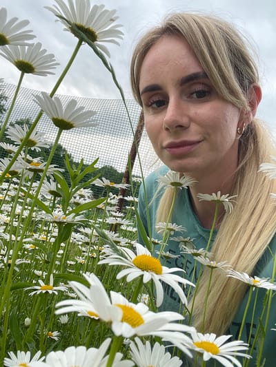 A low-angle, medium shot captures a young Caucasian woman with blonde hair and fair skin in the foreground, looking slightly to her right with a subtle smile. She is wearing a teal t-shirt with a graphic print. Her hair is pulled back into a ponytail, and she has gold hoop earrings. The woman is positioned amidst a field of white daisies with yellow centers, many of which are in focus in front of her. Behind the woman and the flowers, a wire fence is visible, along with some green foliage and the sky, which is overcast with gray clouds. The setting appears to be an outdoor field in Yasnoe, Russia.
