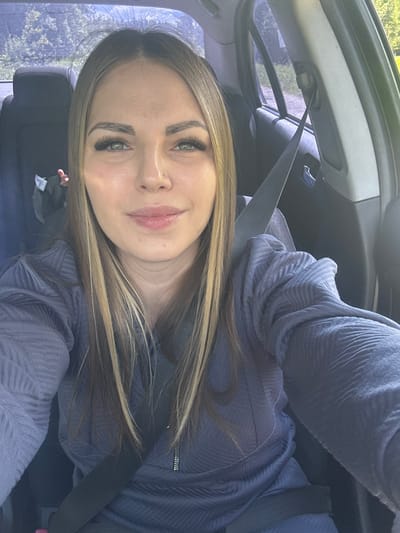A woman with long, light brown and blonde hair and fair skin is shown in a selfie taken inside a car. She is wearing a textured blue zip-up jacket and a seatbelt is fastened across her chest. The background visible through the car windows shows trees and foliage, suggesting a potentially wooded area. The photograph provides no information to suggest this photo was taken in Slobodka, Russia.