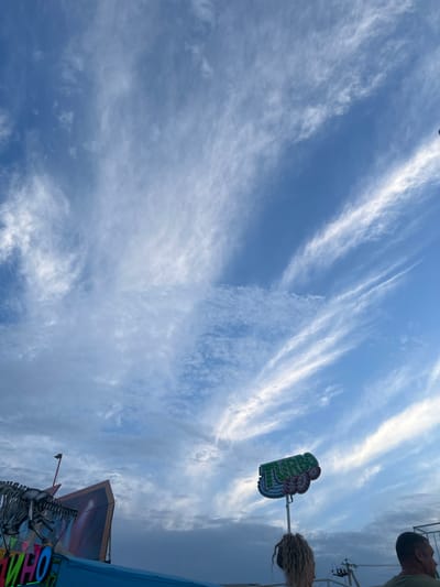 This image was taken in Golubitskaya, Russia, at a seaside amusement park. The sky is a vibrant blue, with thin, wispy clouds streaking across it. In the foreground, a person with curly blonde hair is visible from behind, facing away from the camera. They are standing on what appears to be a ride or attraction, as indicated by a sign overhead with the word "TURBO" in colorful lettering and cartoon-like graphics. To the left of the image, a portion of a dinosaur skeleton structure is visible, part of another amusement ride. The overall scene suggests a lively atmosphere typical of a summer day at an amusement park, with the bright sky and the colorful attractions.