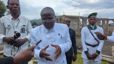 The image depicts a scene outside a partially constructed building in Keshero, Democratic Republic of the Congo, with a prominent figure, likely a politician or religious leader, being interviewed by a reporter with a microphone. The figure is wearing a white long-sleeved shirt with a mandarin collar and multiple buttons, and he has a pen in his pocket. He is standing with his hands clasped together, seemingly listening intently to a question being asked.
To his left, a reporter is holding a microphone and a black handheld device. The reporter is wearing a white shirt with a badge that says "Taxi" and a lanyard with a photo on it. His right hand is holding the microphone, while his left hand is holding the device.
On the right side of the image, two men in white uniforms with green trim and berets are standing, possibly security guards or bodyguards. One is looking at the camera, holding a mobile phone, while the other is looking away from the camera with his hands clasped in front of him.
The background includes a partially constructed building with exposed brick walls and rebar. There are several trees in the distance. The sky is overcast with clouds.