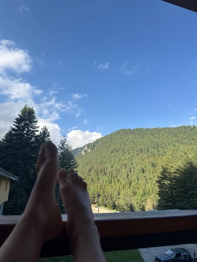 The image shows a person's feet resting on a railing, with a view of a forested mountain and blue sky with clouds in the background. The location is identified as Govedartsi, Bulgaria. The scene appears to be from a balcony or terrace, with a building visible on the left and a dark-colored pickup truck parked on a paved area below on the right. The mountain is covered in dense green pine trees.