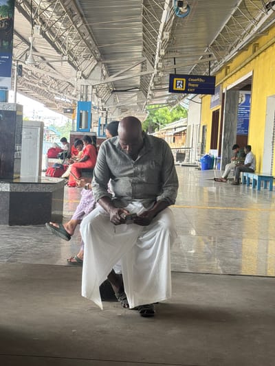 The image depicts a man sitting at a train station in Thanjavur, India. He is bald, wearing a grey shirt and white dhoti, and appears to be looking at a mobile phone in his hands. Several other people are visible in the background, some sitting on benches, and others standing. The environment is an open-air platform with a covered roof supported by metal beams. There are yellow and blue signs on the wall, one of which indicates "EXIT" in multiple languages. The ground is tiled, and there are some potted plants visible in the distance. The overall scene suggests a typical day at a busy Indian railway station.