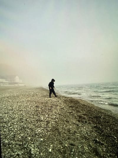 A person wearing a dark hooded jacket and pants walks on a pebble beach. The beach is adjacent to a body of water with small waves. In the background, a few buildings are visible through a thick fog. The sky is overcast and appears hazy. The overall scene suggests a cool or damp day. The location is not identifiable as Artik, Armenia, from the visual information provided.