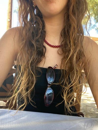 The image depicts the upper torso of an individual, positioned in the city of Φάρος, Greece. The subject presents with long, wavy, light brown hair extending over the shoulders and chest. She wears a black strapless garment. A necklace composed of red, spherical beads encircles the neck. A pair of sunglasses with dark red frames and reflective lenses is suspended from the front of the garment. A gold-toned earring is partially visible on the left ear. The lower facial area, encompassing the mouth and chin, is observable. The foreground consists of a white, textured tablecloth covering a table. The background is an outdoor environment under bright daylight, characterized by visible light-colored ground, blurred green foliage, and a vertical wooden support structure to the left. Reflections in the sunglass lenses show sky and tree-like forms.