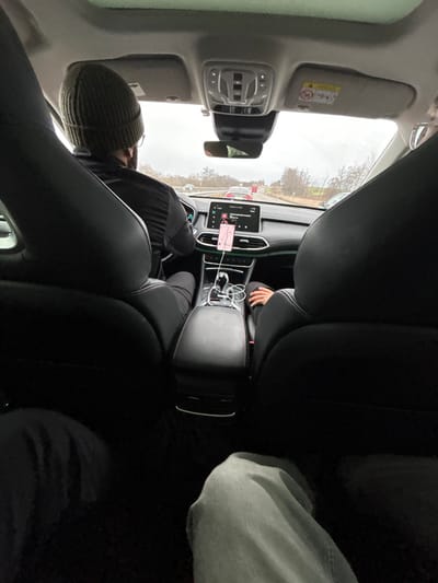 The image displays the interior of an MG HS vehicle from a rear passenger's perspective, looking forward while traveling on a multi-lane road in Steinbourg, France. The environment outside the vehicle is characterized by an overcast sky and roadside trees.

In the front left seat, a male driver is visible from the rear, wearing an olive green knit beanie and glasses, operating the steering wheel. A right hand and forearm, covered by light-colored fabric, are partially visible in the front right passenger seat. The vehicle's dashboard features a central infotainment screen displaying a map and media interface, with a smartphone mounted to its left, also showing a navigation application. The central console between the front seats includes a gear selector, control buttons, and two white USB charging cables. In the foreground, the knees and lower thighs of two individuals occupying the rear seats are partially visible; one is clad in light grey pants, while the other is in dark pants. Multiple vehicles are observed on the road ahead, including red and white cars, indicating traffic flow.