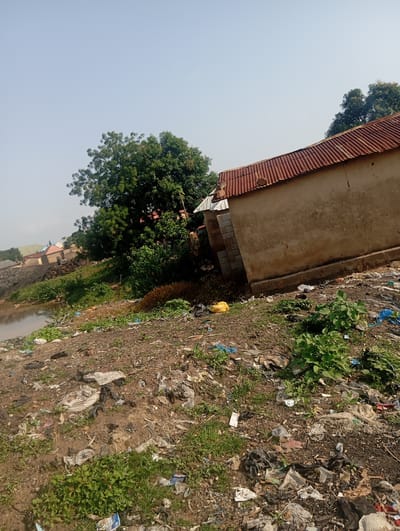 The image depicts a cluttered environment in Gwagwa, Nigeria, with a notable accumulation of refuse. A small, weathered structure with a corrugated metal roof is visible, appearing to be a dwelling or outbuilding, constructed with concrete blocks and mud plaster. The ground surrounding this structure is heavily strewn with various types of trash, including plastic bags, wrappers, and other debris.

In the background, a body of murky water, possibly a pond or river, is present, with its banks also showing signs of litter. Adjacent to the water and the immediate foreground, there is a growth of green vegetation, including trees and smaller plants, some of which are partially obscured by the refuse. Further in the distance, several other buildings with colorful roofs are scattered across the landscape, suggesting a residential area. The sky is clear and pale blue, indicating daytime. No people are visible in the frame. The overall impression is one of environmental neglect and inadequate waste management.
