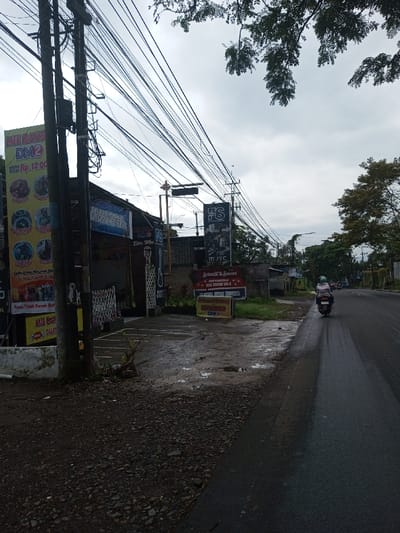 The image depicts an outdoor street scene under an overcast sky, with visible evidence of recent rainfall on the asphalt road.
On the left side of the frame, multiple utility poles are present, heavily laden with a dense network of overhead electrical and communication cables. Attached to one prominent pole is a large vertical banner advertising "MIE BAKSO DM2" with images of various food items and a price of "Rp. 12.000." Another banner is partially visible below it, also mentioning "MIE BAKSO." Behind these poles and banners, a building entrance is visible, adorned with a horizontal banner that reads "SELAMAT DATANG DI FS STUDIO MUSIK" (Welcome to FS Music Studio). A white and multi-colored metal barrier or fence stands in front of this entrance. The ground in the immediate foreground on the left is unpaved, consisting of dirt and gravel. Further right on the left side, another brown and white sign with the "FS" logo and text related to "MUSIC STUDIO" is positioned near the road, alongside a smaller red and white banner displaying "SELAMAT & SUKSES KEGIATAN FS MUSIC STUDIO DM2 MIE BAKSO DM2 INDONESIA."
The right side of the image features a wet, dark asphalt road extending into the distance. A single individual, wearing a light-colored garment and a green helmet, is operating a black scooter, moving away from the viewer along the right lane. Other vehicles are faintly discernible in the far distance on the road. Trees and green foliage line the roadside further along, and additional utility poles with overhead wires are visible. The sky is uniformly gray and cloudy. The linguistic content on the banners (Indonesian language) and currency notation (Rp. for Rupiah) indicate the location is consistent with Indonesia, not Kamji Na-Yekha, Bhutan.