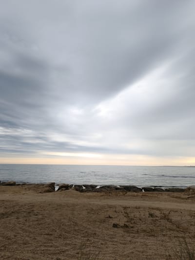 The image depicts a seascape with a cloudy sky over a calm body of water. In the foreground, there is a sandy beach with scattered rocks and dry vegetation. The horizon line separates the water from the sky, which is filled with various cloud formations, some appearing light and wispy, while others are darker and more dense. A faint yellowish-orange glow is visible near the horizon, suggesting either sunrise or sunset. The water is a muted blue-grey, with gentle ripples on its surface. Along the shoreline, a collection of larger, dark rocks forms a natural barrier between the sand and the sea. The overall mood of the image is serene and somewhat overcast. This scene is located on the coast of the Caspian Sea in the Sabunchu Raion of Azerbaijan.