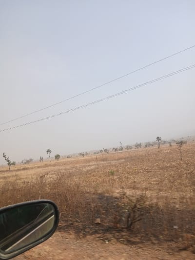 This image captures a wide-angle, exterior view of a dry, open landscape in Bokana, Nigeria, as observed from a moving vehicle. The lower left of the frame is dominated by a dark vehicle side-view mirror, which features a prominent diagonal crack and reflects a portion of a road line and interior vehicle elements. The terrain consists of vast expanses of reddish-brown, dry earth and sparse, desiccated vegetation, indicating a dry season. Scattered individual trees, including some taller palm-like specimens and smaller shrubs, are distributed across the midground and background, extending towards a hazy horizon. Darker patches consistent with scorched earth are visible in the foreground and midground. The sky above is a pale, desaturated blue-grey, and two parallel utility lines, appearing as dotted traces, diagonally traverse the upper-middle section of the frame. No human or animal subjects are present.
