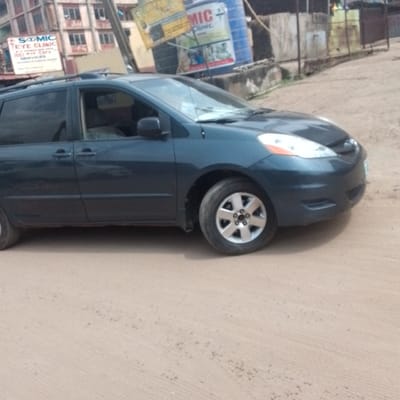 A dark blue Toyota Sienna minivan is parked on a dirt road in Awada, Nigeria. Behind the minivan are buildings and signage, including one for "Scomic Eye Clinic" and another with "MIC Clinic" printed on a water tank. The general environment suggests a commercial or residential street in an urban area.