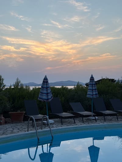 This image depicts an outdoor swimming pool area at dusk, with a body of water and distant hills visible in the background. The sky is filled with wispy clouds illuminated by the setting sun, casting warm hues of orange and pink. Several empty lounge chairs are arranged along the poolside, some with umbrellas partially open. A terra cotta pot and a decorative lantern are visible on the stone-paved area behind the chairs. The pool water is a vibrant blue, reflecting the sky and some of the surrounding elements. The overall scene suggests a tranquil, resort-like setting, possibly located in a coastal area near Alghero, Italy, given the presence of water and hilly terrain suggestive of the Sardinian landscape. No people are present in the image.