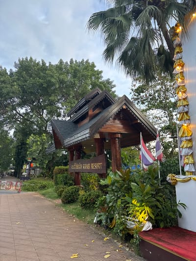 The image depicts an outdoor scene in Ban Klong Haeng, Thailand, featuring the entrance to "Krabi Resort". A wooden sign with Thai script and the English words "KRABI RESORT" is prominently displayed. The resort's architecture features a traditional Thai-style roof with intricate detailing. Two Thai flags are planted near the entrance, one red, white, and blue, and another with similar colors. Lush green vegetation surrounds the entrance, with a paved walkway leading to it. In the background, palm trees and other trees are visible, under a cloudy sky. Decorative yellow and white fabric garlands with lights are draped along a white wall on the right side of the image. The scene suggests a welcoming entrance to a tropical resort.