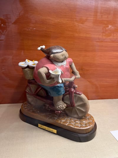 A ceramic sculpture depicts a child riding a bicycle. The figure wears a pink top, blue shorts, and a dark cap, holding white flowers. The bicycle has reddish-brown details. The sculpture sits on a textured, brown base with a dark brown trim. A gold label is affixed to the side of the base. The sculpture is positioned against a light brown wall and a darker wooden surface.
